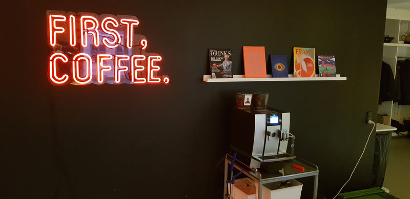 Rødt neonskilt med FIRST, COFFEE. ved en kaffemaskine på et kontor