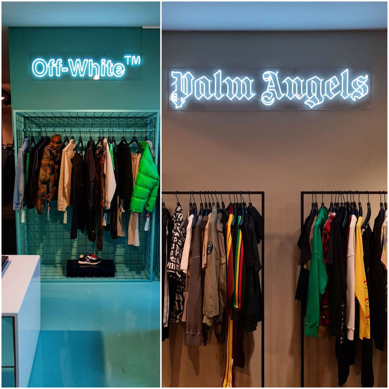 Hvidt neonskilt med Palm Angels-logoet over tøjstativer i en butik