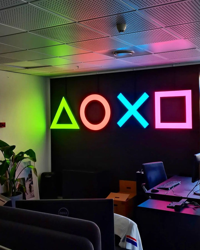 PlayStations fire symboler, trekant, cirkel, kryds og firkant, som LED-neonskilte i grøn, koral, turkis og pink på en mørk kontorvæg